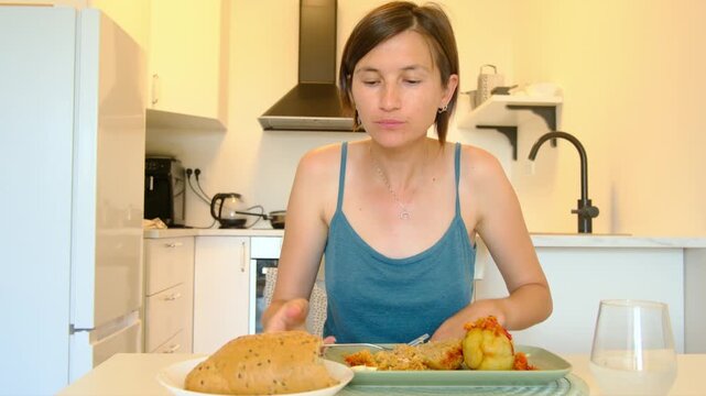 Woman sits at kitchen table eating stuffed paprika, enjoying homemade Balkan cuisine, turning meal into cultural moment shared in quiet domestic space.