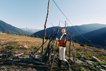 Naklejka premium Woman in sweater and boots standing by a rustic fence in a rural field with mountains in the background, countryside landscape and clear sky for outdoor lifestyle travel