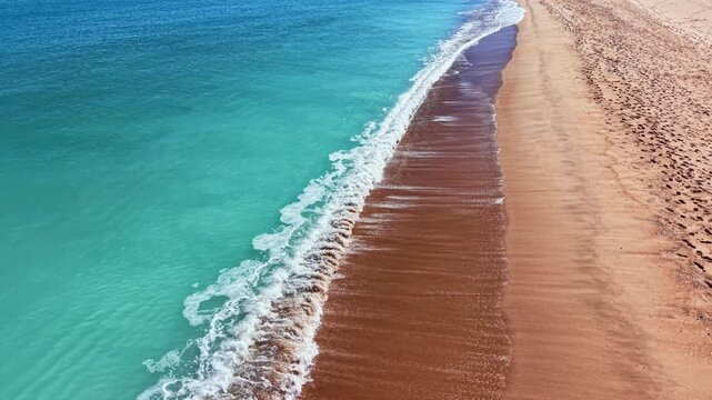 Footprints are visible in the sand as waves gently hit the shore on a sunny summer day. The clear water meets the golden beach, creating a calm scene.
