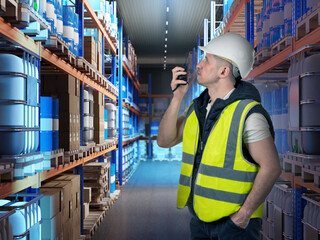 Warehouse, logistics center. Worker in hard hat uses handheld radio while checking shelves with...