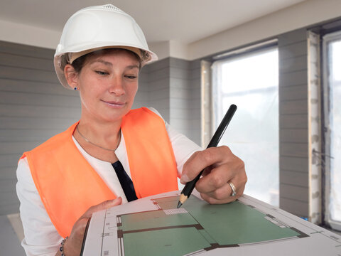 Construction, architectural supervision. Female engineer in hard hat reviews building plan inside unfinished house, marking layout details, quality control, renovation planning, or site inspection.