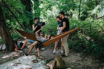 Fototapeta premium Friends relax in a hammock under green trees in a sunny forest park. The scene captures a casual outdoor hangout with smiles and playful energy.