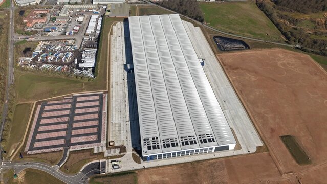 Aerial view of an immense warehouse with a silver roof casting long shadows over the brown earth, contrasting with the neatly arranged parking lot, Doncaster, United Kingdom.