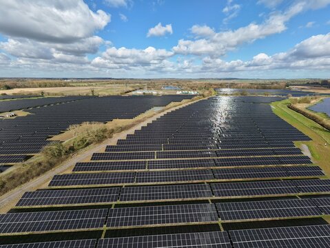 Aerial view of rows of solar panels reflecting the sky, interspersed with green fields and water bodies under a sky with fluffy clouds, Retford, United Kingdom.