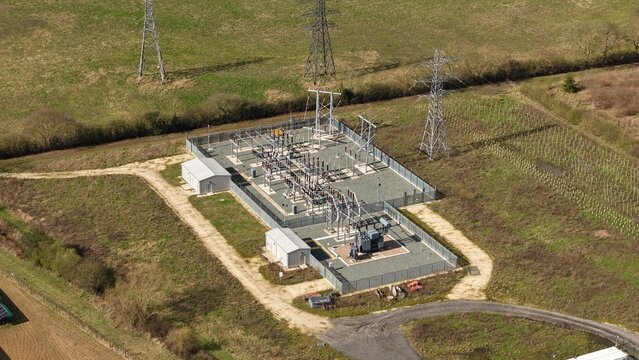 Aerial view of an electrical substation standing stark against the green landscape, power lines reaching into the bright sky, Retford, United Kingdom.