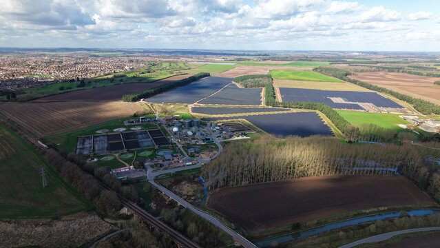Aerial view of sun-kissed wastewater treatment plant nestled among patchwork fields, where deep blues meet earthy browns under a vast, cloud-strewn sky, Worksop, United Kingdom.