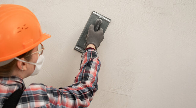 Builder woman in hardhat and protective mask sanding and smoothing walls after plastering for leveling getting it ready for painting during home renovation and construction work.