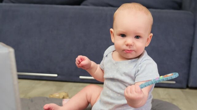 Adorable baby boy sitting on the floor, curiously exploring a teether toy shaped like a remote control while looking at the camera, developing fine motor skills and hand-eye coordination