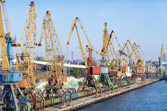 Line of tall portal cranes stands on concrete pier near blue sea water. Freight train cars move along tracks for bulk cargo loading under clear sky. Industrial port infrastructure in daylight.