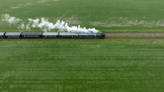 Aerial view of a vintage steam train chugging through vibrant green fields, its white smoke contrasting against the lush landscape, Benningbroek, Noord-Holland, Netherlands.