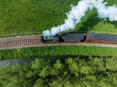 Aerial view of a vintage steam train chugging along the tracks, plumes of white smoke billowing against the lush green fields, Benningbroek, Noord-Holland, Netherlands.