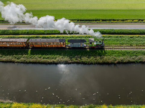 Aerial view of a vintage steam train chugs along the tracks, contrasting with the bright green fields and a dark canal, Benningbroek, Noord-Holland, Netherlands.