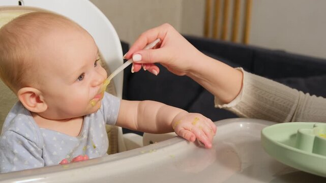 Mother's hand feeding a little caucasian baby boy with vegetable puree from a spoon. Adorable infant sitting in a high chair at home eating his first solid food during weaning