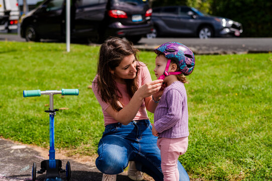 Italian mum securing toddler daughters helmet outdoors on paved path