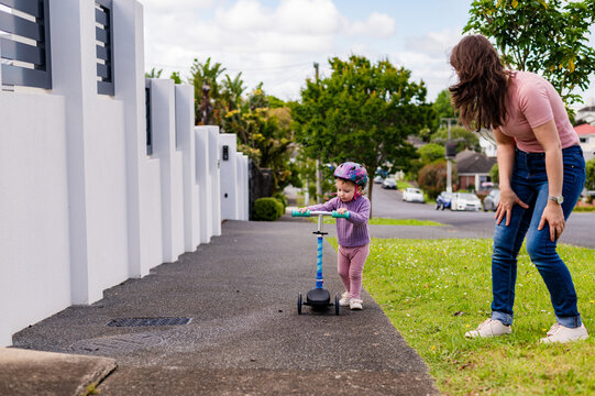 Mum guiding toddler daughter in riding her scooter outdoors