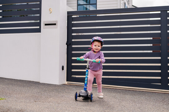 Toddler girl riding on her scooter on paved path with her helmet on outside home