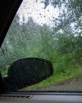 View from inside a car through a rain-covered window with visible side mirror. Wet glass with raindrops and blurred green forest landscape along a rural road during rainy weather. Atmospheric travel