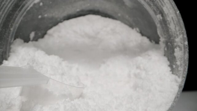 Inside the jar with white powder, a measuring spoon is filled with creatine, a sports supplement. Dolly slider extreme close-up.