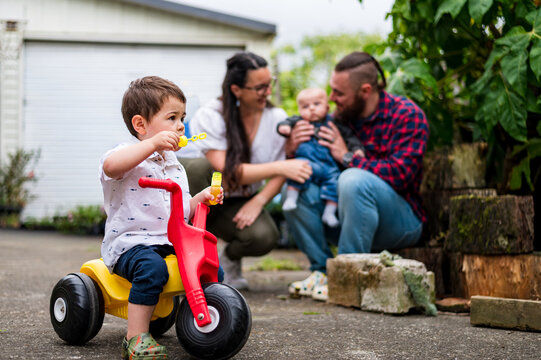 Toddler boy on his trike toy blowing bubbles in the backyard with parent in the background