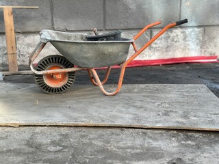 A weathered metal wheelbarrow with an orange frame and worn wheel on a concrete surface at a...