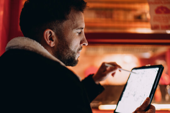 Close up of man drawing on tablet with stylus at night, male designer focused on digital artwork, 5G wireless internet technology, remote work, digital nomad life, authentic representation, profile.