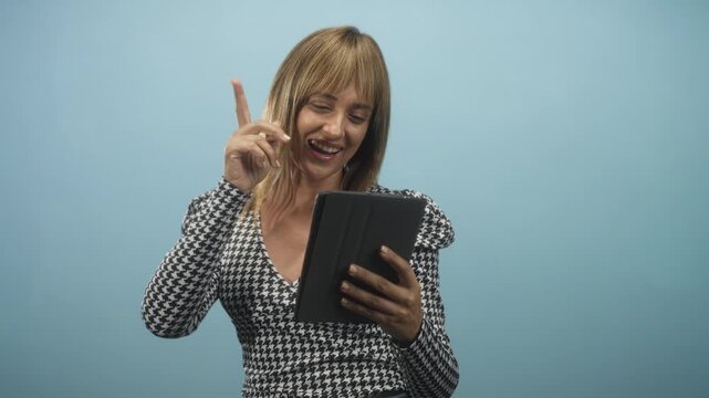 Woman holding tablet, smiling broadly with right hand clenched in a fist pump gesture, wearing houndstooth top against blue studio backdrop; joy celebration.