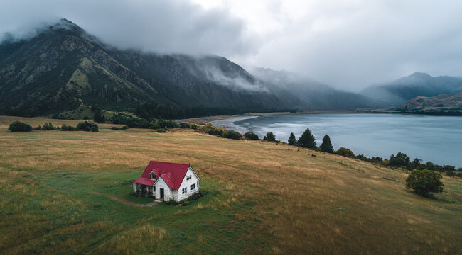 A solitary red-roof house sits in a grassy field with a lake and misty mountains in the background, captured in cinematic style under a moody sky.  