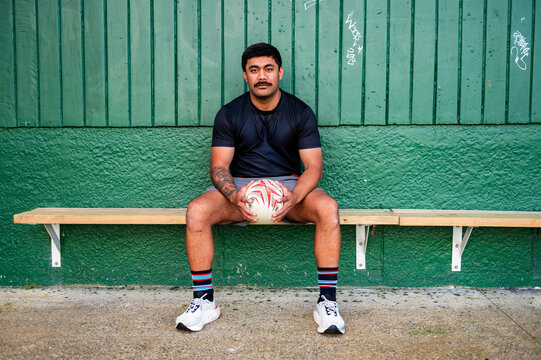 Samoan man sitting on wooden bench holding a rugby ball in waiting area