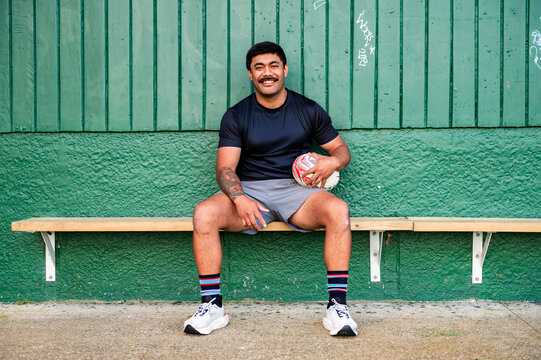 Samoan man sitting on wooden bench holding a rugby ball in waiting area