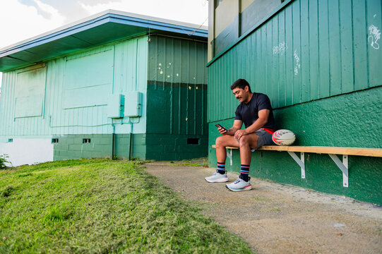 Samoan rugby player sitting on wooden bench holding a phone