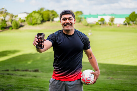 Smiling man holds a phone and a football, enjoying practice at a sunny field