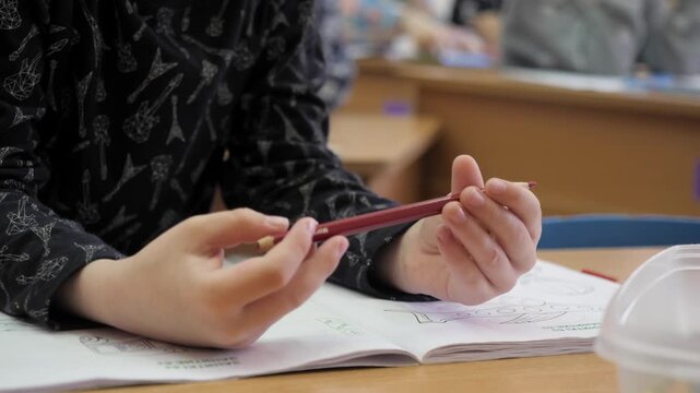 small hands holding wooden red pencil over open coloring book pages on classroom desk during creative art lesson inside bright preschool kindergarten facility.