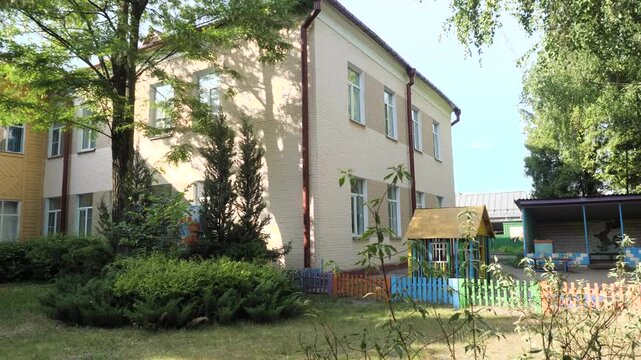 Exterior view showcasing length of two story light beige brick kindergarten building with white painted lower section and red drainage pipes beside lush green lawn