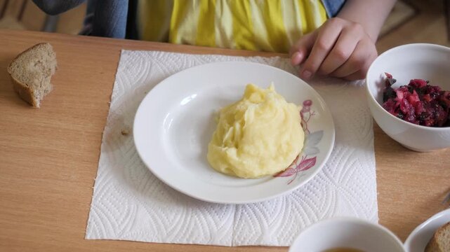 Young child sitting at wooden table eating simple lunch consisting of mashed potatoes piece of boiled sausage slice of rye bread beetroot salad tea in kindergarten canteen.