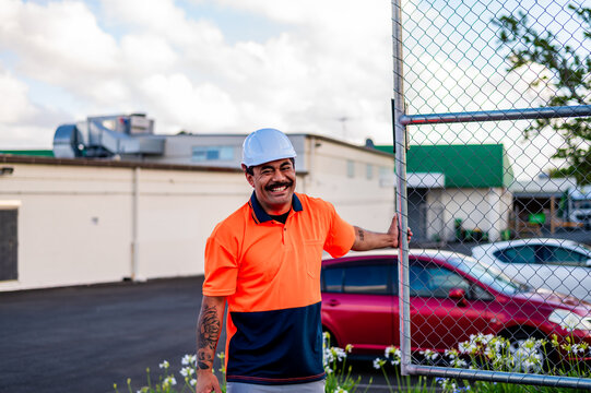 Man in work attire stands by a fence, smiling in a vibrant city setting under a bright sky