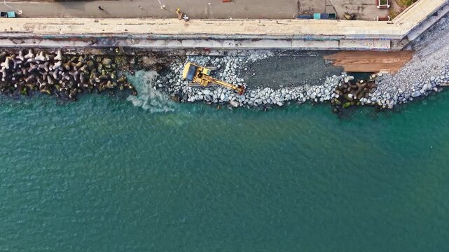 A machine moves rocks along the shore near water while workers are present. The construction process is ongoing in a coastal setting during the day.