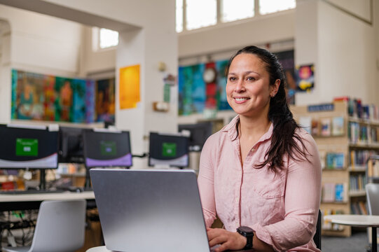 Woman sits at table in library using laptop, focused on task with bookshelves in background