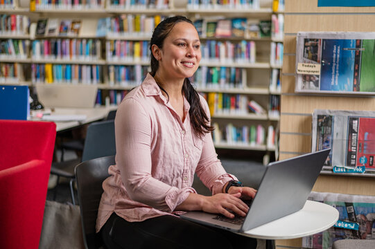 Woman sits at a table in a library using a laptop while smiling at the screen