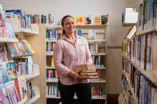 Woman stands between shelves, holding books in a library during the day in New Zealand