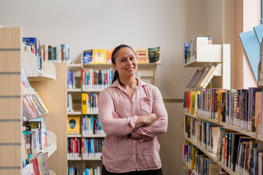 A woman stands smiling in a library surrounded by organised shelves and books with arms crossed