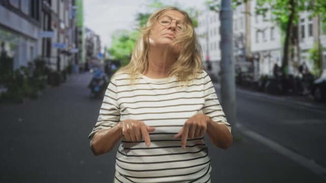 Senior woman pointing both index fingers down at pavement on a city street wearing striped shirt; quiet contemplation.