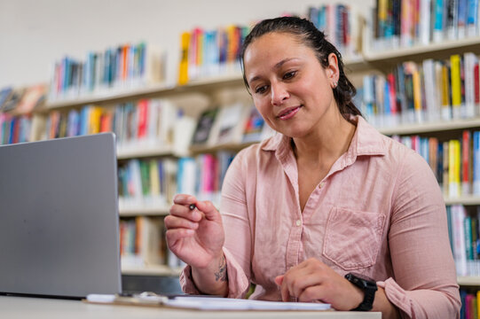 Woman sits at a table in a library using a laptop and writing notes