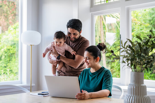 Indian dad holding baby girl beside mum working on a laptop at a wooden table in a bright room