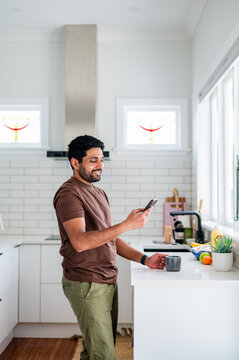 Indian man standing scrolling through his phone while holding a cup near the kitchen counter