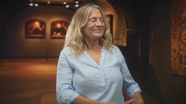 Senior woman, blonde, gestures with hands presenting a framed painting in a warm lit museum gallery with arched ceiling and rope barrier, smiling and explaining; contemplation appreciation.