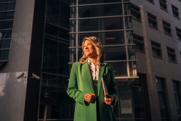 Fototapeta premium Confident businesswoman smiling holding laptop outside corporate building