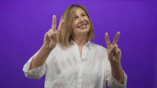 Middle age woman in white eyelet blouse holds both hands up showing peace signs in studio setting; happiness.