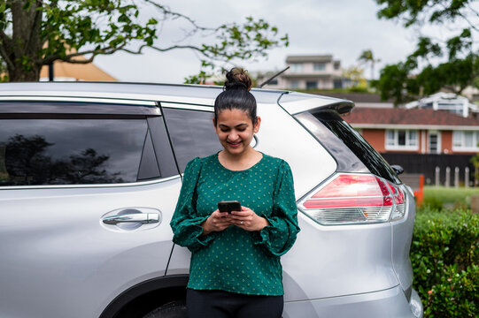 Woman standing outside a silver car and looking at her smartphone in a suburban location