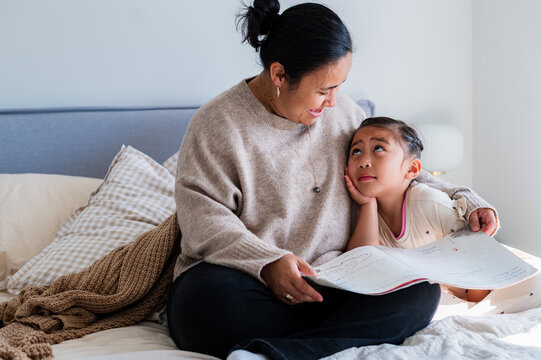 Young girl looking up to mum reading a story while seated on the bed