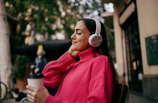 Cheerful girl wearing white headphones enjoying audio content at sidewalk terrace, happy young woman with coffee cup smiling with closed eyes, mindfulness and stress relief in urban environment.